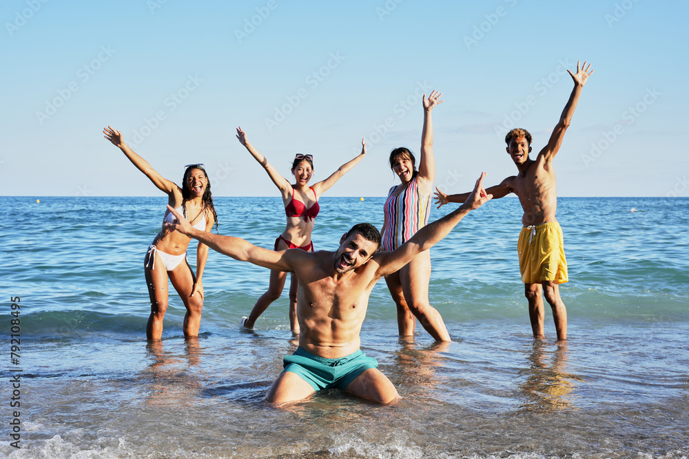 Portrait of a group of happy young people in swimming costumes posing for a photo on the shore of the beach. Diverse friends smiling and making playful gestures raising arms looking at camera Portrait of a group of happy young people in swimming costumes posing for a photo on the shore of the beach. Diverse friends smiling and making playful gestures raising arms looking at camera