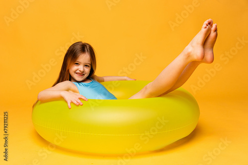 Happy funny little girl dressed in a blue swimsuit lying on an inflatable color ring on yellow background. Summer vacation concept. Beach time. Copy space for ad.