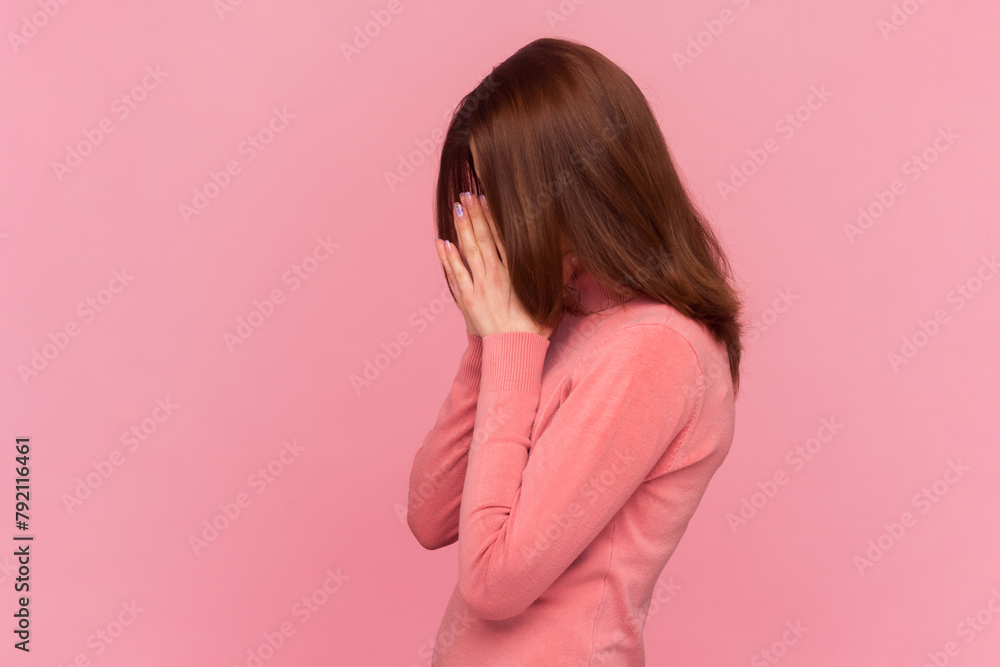 Side view portrait of sad desperate woman with brown hair hiding her face and crying having serious troubles, being in bad mood, wearing rose turtleneck. Indoor studio shot isolated on pink background Side view portrait of sad desperate woman with brown hair hiding her face and crying having serious troubles, being in bad mood, wearing rose turtleneck. Indoor studio shot isolated on pink background