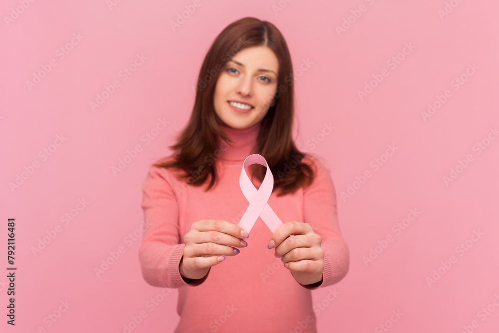 Female health. Woman with brown hair holding pink ribbon, concept of breast cancer awareness, oncological disease prevention, wearing rose turtleneck. Indoor studio shot isolated on pink background Female health. Woman with brown hair holding pink ribbon, concept of breast cancer awareness, oncological disease prevention, wearing rose turtleneck. Indoor studio shot isolated on pink background