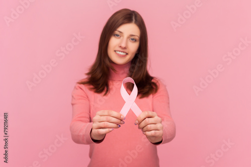 Female health. Woman with brown hair holding pink ribbon, concept of breast cancer awareness, oncological disease prevention, wearing rose turtleneck. Indoor studio shot isolated on pink background photo