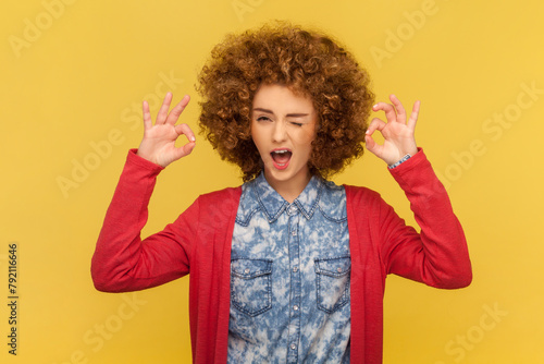 Portrait of playful cheerful woman with Afro hairstyle winking to camera, showing okay gesture with both hands, saying all is ok. Indoor studio shot isolated on yellow background.