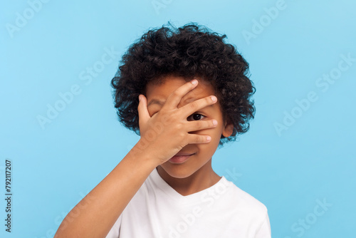 Portrait of shy curious little boy with curly hair looking at camera trough his fingers spying sees something shameful. Indoor studio shot isolated on blue background.