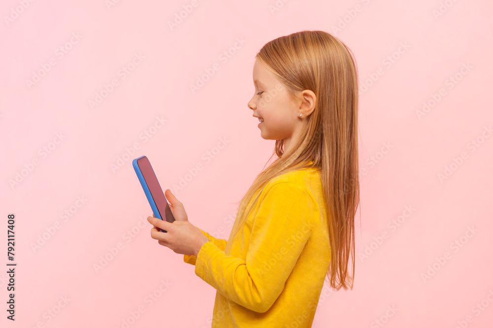 Naklejka premium Side view portrait of adorable cute blonde little girl using cell phone with happy expression, playing games, enjoying, wearing yellow jumper. Indoor studio shot isolated on pink background.