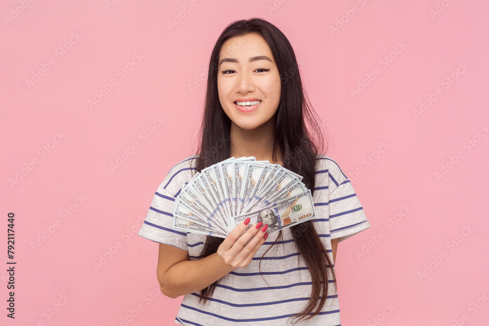 Portrait of satisfied pride rich woman with long brunette hair holding fan of dollar banknotes, wearing striped T-shirt. Indoor studio shot isolated on pink background. Portrait of satisfied pride rich woman with long brunette hair holding fan of dollar banknotes, wearing striped T-shirt. Indoor studio shot isolated on pink background.