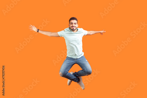 Full length portrait of jumping young bearded man wearing T-shirt feeling inspiration rejoicing expressing happiness flying in air. Indoor studio shot isolated on orange background.