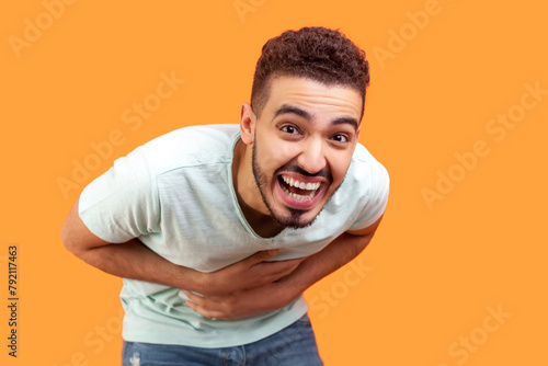 Portrait of happy joyful cheerful young bearded man wearing T-shirt laughing out loud holding belly hearing funny joke. Indoor studio shot isolated on orange background.