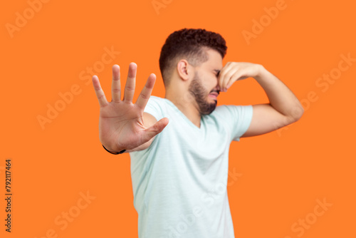 Portrait of displeased young bearded man wearing T-shirt pinching his nose turning away feeling disgust bad smell. Indoor studio shot isolated on orange background. photo