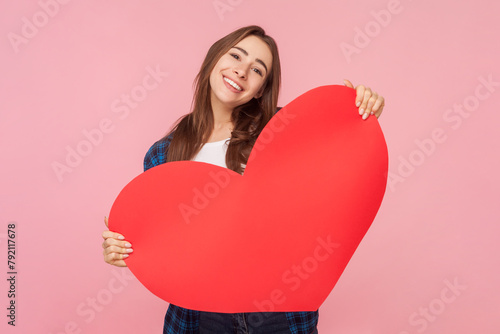 Portrait of smiling good looking brown haired woman holding big red heart looking at camera with positive expression, wearing checkered shirt. Indoor studio shot isolated on pink background. © khosrork