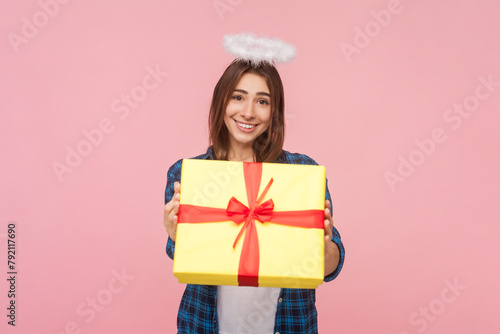 Portrait of cute smiling angelic brown haired woman with nimb over head giving present box, congratulating, offering gift, wearing checkered shirt. Indoor studio shot isolated on pink background.