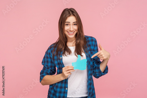 Portrait of cheerful brown haired woman showing pointing paper thumb up asking to appreciate her blog content, wearing checkered shirt. Indoor studio shot isolated on pink background.