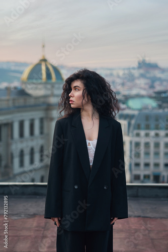 Attractive young curly brunette woman in a black jacket and black pants stands on a rooftop against a historic building during a magical sunset.
