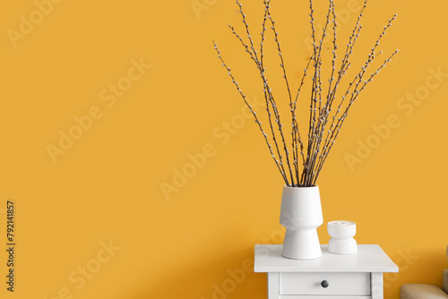 Vase with willow branches on table near yellow wall