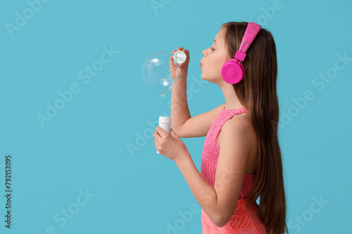 Cute girl in headphones blowing soap bubbles on blue background