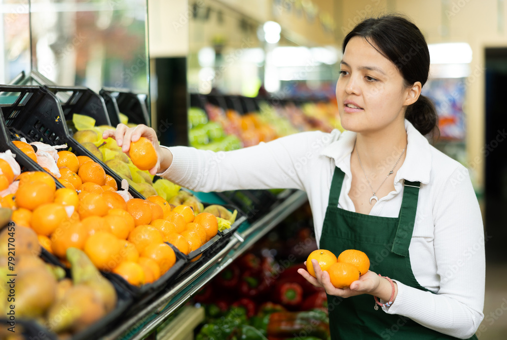 Young woman seller in apron lays out tangerines in supermarket