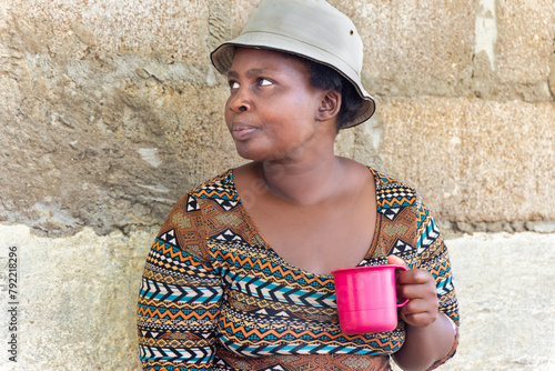 young african chubby woman with a hat holding a mug in the poor township, informal settlement