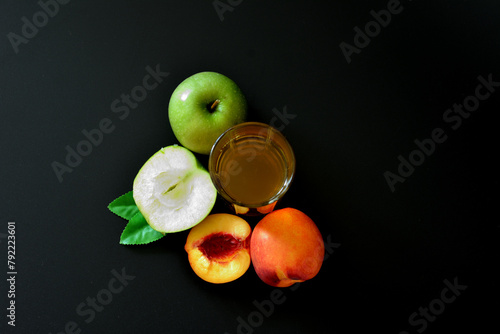A tall glass of fruit juice on a black background, next to pieces of a ripe peach and a green apple.