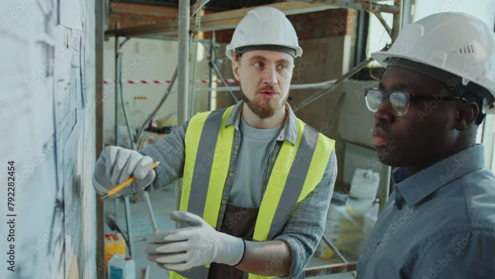 Caucasian construction site worker in hardhat, gloves and safety vest pointing at architectural drawings on the wall and talking with African American engineer Caucasian construction site worker in hardhat, gloves and safety vest pointing at architectural drawings on the wall and talking with African American engineer