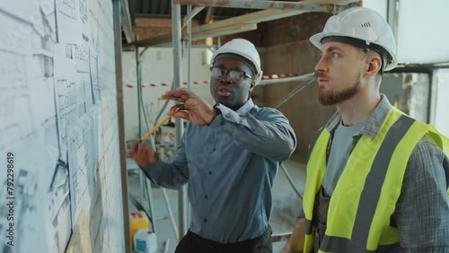 African American male architect in hardhat and formal shirt pointing at floor plans on wall and giving instructions to Caucasian building contractor at construction site