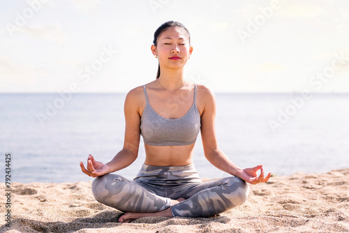 young asian woman doing meditation at beach sitting with legs crossed, concept of mental relaxation and healthy lifestyle