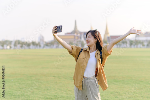 Traveler asian woman in her 30s making a livestream or selfie on smartphone while explores Wat Phra Kaew emerald Buddha. Share the wonders of Thai heritage through her journey.
