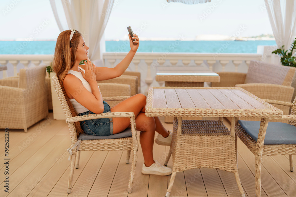 A woman is sitting at a table with a cell phone in her hand. She is taking a picture of the ocean.