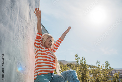 A woman is sitting on a railing with her arms raised in the air. The sun is shining brightly, creating a warm and inviting atmosphere.