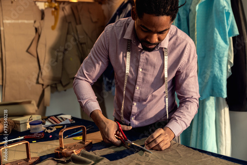 Male fashion tailor working on the suit design in his workshop cutting fabric with scissors.