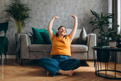 Obraz na płótnie Calm Smiling girl relaxing at home, she is listening to music, using a smartphone and wearing white headphones. Young 30s woman hearing enjoying good audio sound lounge near couch at home, full body w salonie
