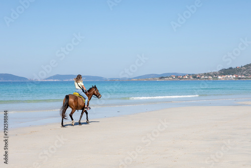 A female equestrian rides a brown horse along the shoreline with clear blue waters and a mountainous backdrop, capturing a moment of peaceful coexistence with nature