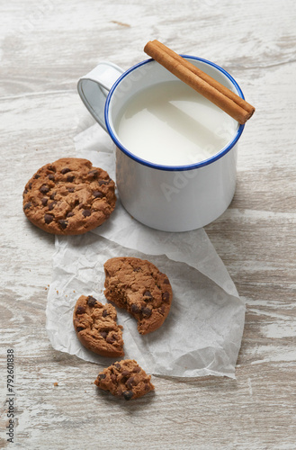 A rustic mug of milk garnished with a cinnamon stick beside freshly baked chocolate chip cookies on a weathered wooden table photo