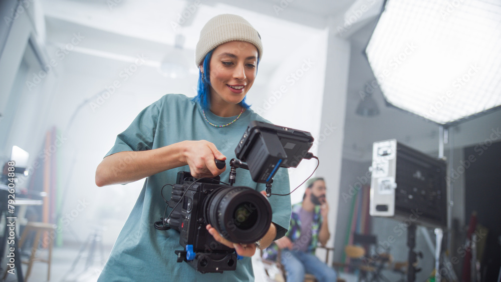 Young Female Cinematographer With Blue Hair Adjusts Professional Camera In Studio, Showcasing Teamwork In Creative Film Production Environment, With Colleagues Collaborating In Background. Young Female Cinematographer With Blue Hair Adjusts Professional Camera In Studio, Showcasing Teamwork In Creative Film Production Environment, With Colleagues Collaborating In Background.