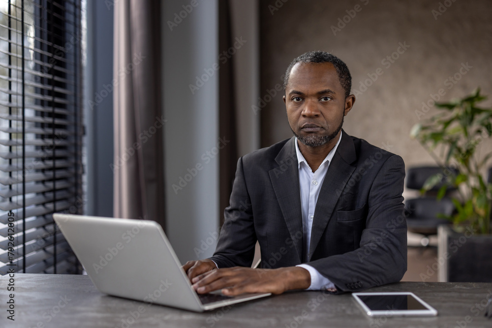 Focused African American businessman in a dark suit works from his laptop in a well-lit modern office, exuding confidence and expertise. Focused African American businessman in a dark suit works from his laptop in a well-lit modern office, exuding confidence and expertise.