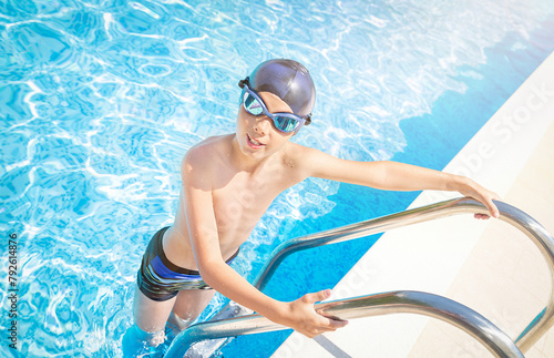 Child (boy) in cap and sport goggles on ladder handrails (stairs) in to the swimming pool, smiling after swimming in summer day. Kid enjoying water. Healthy lifestyle. Copy space.