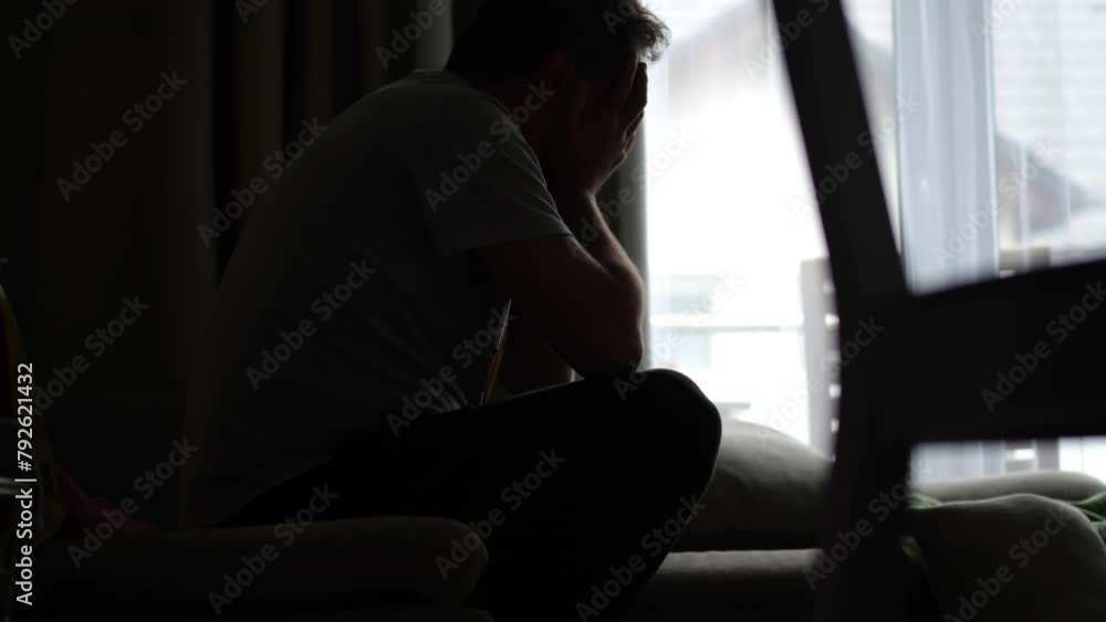 A man in despair rocks on a sofa with his face in his hands, in a dark room with light from a window and an empty alcohol bottle nearby. A man in despair rocks on a sofa with his face in his hands, in a dark room with light from a window and an empty alcohol bottle nearby.