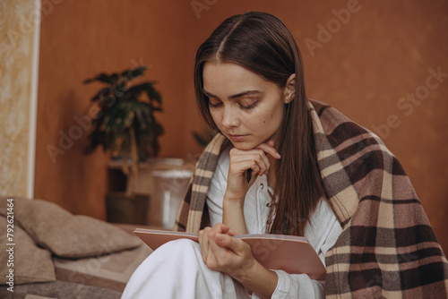 Thoughtful young woman wrapped in cozy plaid reading diary on bed at home 