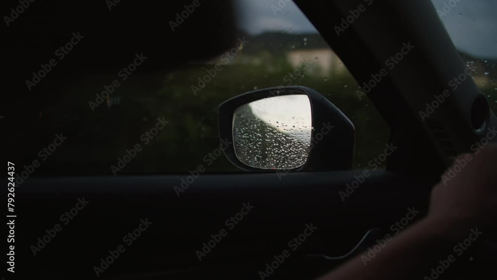 View from inside the car, side mirror, with raindrops, partial view of the driver's hand on the steering wheel and a vague reflection of the building and trees in the mirror View from inside the car, side mirror, with raindrops, partial view of the driver's hand on the steering wheel and a vague reflection of the building and trees in the mirror