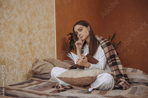 Thoughtful woman in cozy plaid reading book on bed at home 