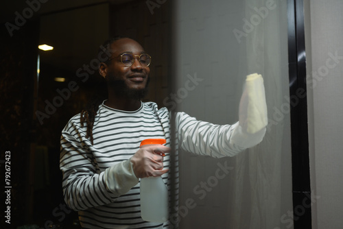 Smiling man in rubber gloves cleaning mirror with rag and spray in a modern bathroom