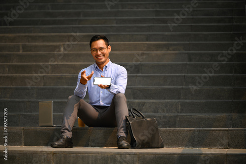 Cheerful businessman sitting on stairs of office building and showing smartphone with blank screen