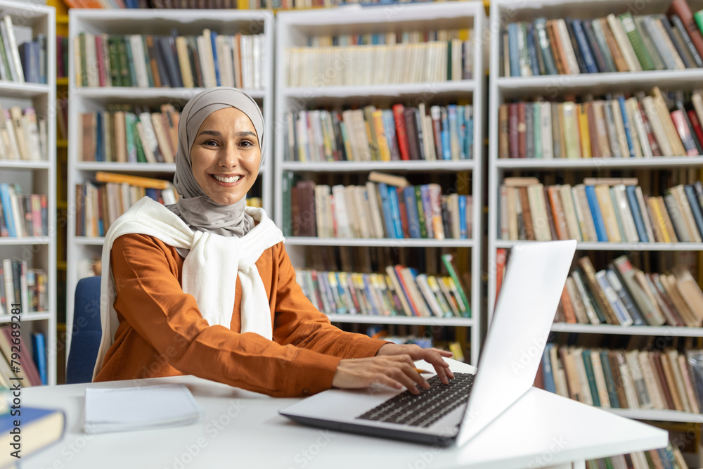 A cheerful Muslim woman in hijab sitting at a desk in a library ...