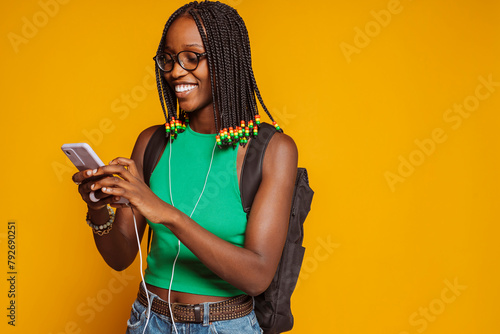 Young happy smiling african feamle student, wears green top, braids, eyeglasses and backpack, listening to music on headphones and smartphone isolated over yellow background at studio. photo