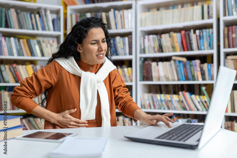 A young female student in a library clutching her stomach in pain, struggling with a stomach ache as she uses her laptop. A young female student in a library clutching her stomach in pain, struggling with a stomach ache as she uses her laptop.