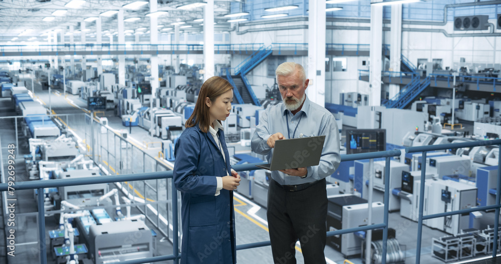 Factory Managers Working on Laptop at a Modern Manufacture with Autonomous Robot Hands. Female and Male Specialists Monitoring Technical Data on a Computer, Analyzing Reports and Planning Updates Factory Managers Working on Laptop at a Modern Manufacture with Autonomous Robot Hands. Female and Male Specialists Monitoring Technical Data on a Computer, Analyzing Reports and Planning Updates