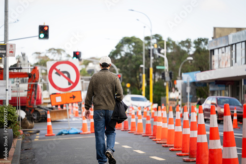 Man walking on the road with orange traffic cones diverting the traffic. Bus stop blocked by the traffic cones. Roadworks in Auckland.