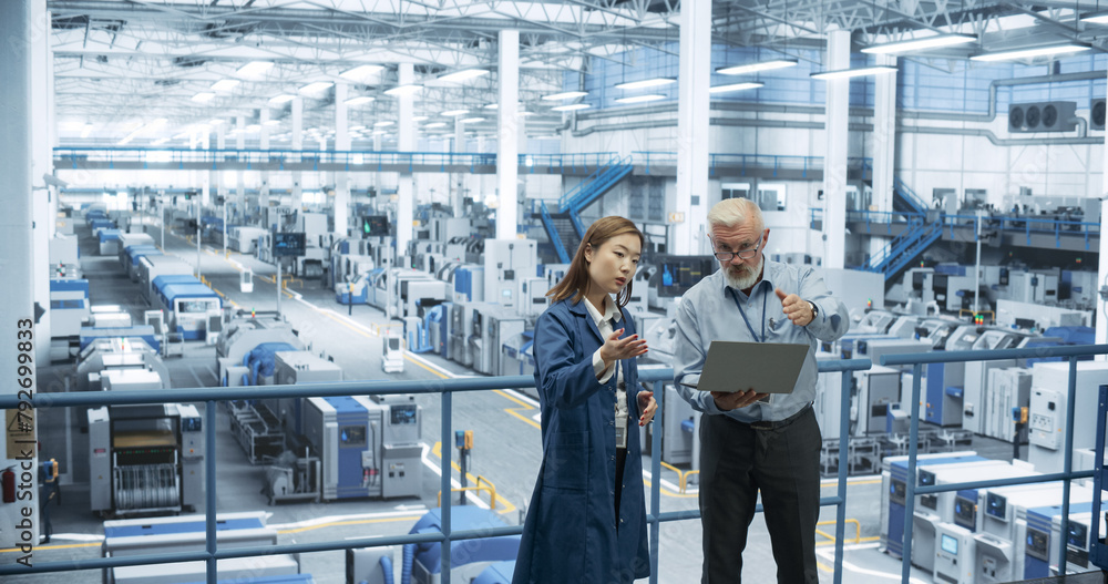 Multiethnic Male and Female Colleagues Standing in a Manufacturing Complex, Using Laptop Computer and Discussing Production on Modern Automated Electronic Devices and Gadgets Multiethnic Male and Female Colleagues Standing in a Manufacturing Complex, Using Laptop Computer and Discussing Production on Modern Automated Electronic Devices and Gadgets
