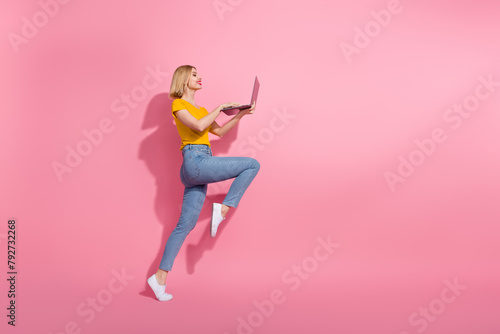 Full length photo of cute excited lady dressed yellow t-shirt jumping typing emails device empty space isolated pink color background