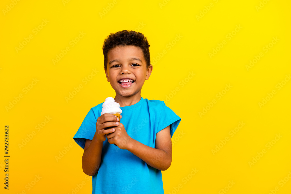 Portrait of pleasant little schoolboy with afro hair wear blue t-shirt holding ice cream in hands isolated on vivid yellow background Portrait of pleasant little schoolboy with afro hair wear blue t-shirt holding ice cream in hands isolated on vivid yellow background
