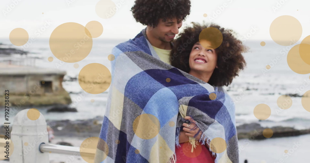 Fototapeta premium Image of light spots over biracial couple covered in blanket embracing on beach