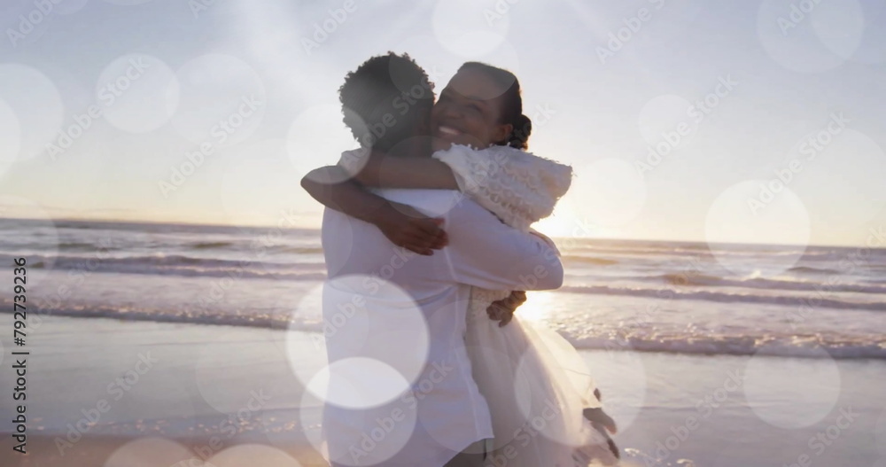 Naklejka premium Image of light spots over happy african american bride and groom embracing on beach at wedding